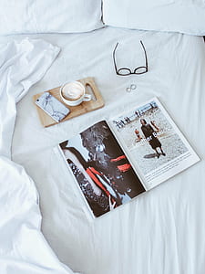 white magazine, coffee in cup, black eyeglasses on bed thumbnail