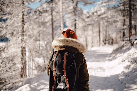 person wearing parka jacket standing on snow covered road surrounded by trees during daytime thumbnail