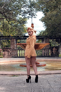 woman wearing brown coat standing beside water fountain near trees under blue sky at daytime thumbnail