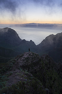 man standing on mountain cliff overlooking body of water under grey cloudy sky during daytime thumbnail