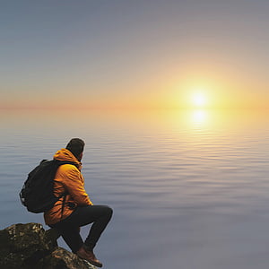 sitting man on rock formation wearing yellow pullover hoodie and black pants in front of body of water thumbnail