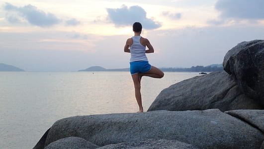 woman wearing white racerback tank top with blue shorts standing on rock near body of water during daytime thumbnail