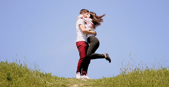 girl hugging boy with raising left foot on air surrounded by green grass in the hill under blue sky during daytime thumbnail