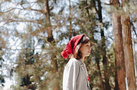 woman wearing red knit cap and gray jacket standing near brown and green high trees under white sky during daytime thumbnail