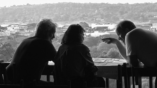 grayscale photo of three people leaning on wood rail talking thumbnail