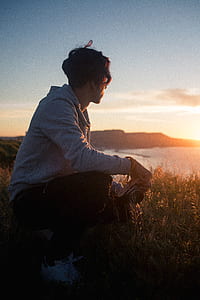man sitting on green grass holding camera while watching sunset thumbnail