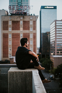 man in black crew-neck shirt and black pants sitting on edge of a high surface during daytime thumbnail