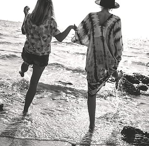 Grayscale Photograph of 2 Women Holding Hands While Walking on Sea Shore thumbnail