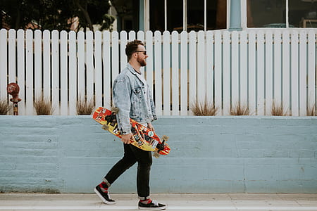 man in blue denim button-up jacket and black pants holding multicolored longboard during daytime thumbnail