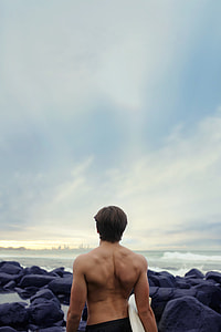 man standing on rocky beach shore holding white surfboard thumbnail