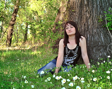 woman wearing black tank top and blue jeans sitting on grass near tree thumbnail