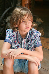 smiling boy wearing white, red, and blue plaid sport shirt and blue denim shorts sitting on brown wooden floor thumbnail