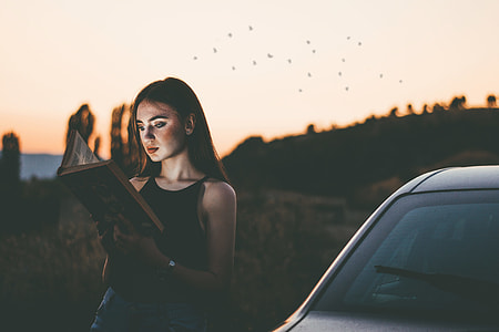 photo of a woman reading book while leaning on car during daytimne thumbnail