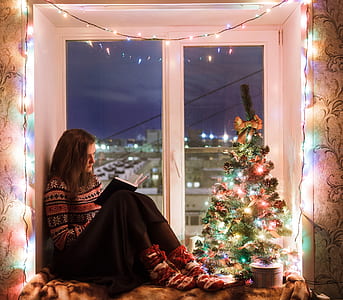 woman in brown and black sweater and black bottoms with socks sitting besides white wooden framed clear glass window pane with string light in front of mini Christmas tree reading book thumbnail