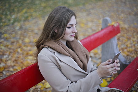 Woman in Beige Coat Holding Smartphone Sitting on Bench thumbnail