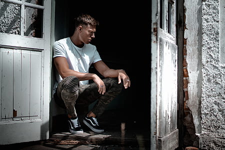 man sitting beside gray wooden wall in front of opened door during daytime photo thumbnail