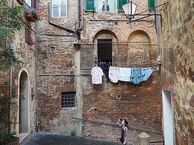woman in gray cardigan holding camera and taking photo of floral arrangement on 3rd storey window of brick residential building during daytime thumbnail