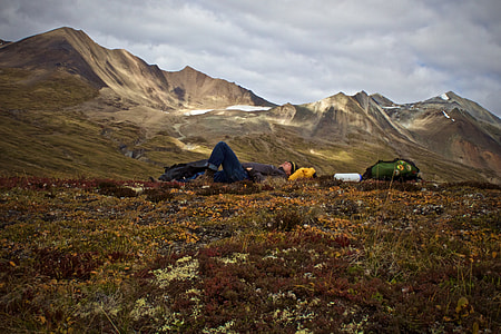 man lying on hill beside green hiking backpack near mountains under white and blue cloudy skies thumbnail
