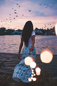 woman standing in front of body of water looking at flying birds during sunset thumbnail