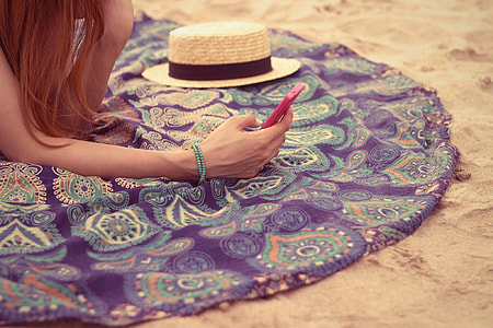 person holding red android phone and brown and black sun hat sitting in the white sand thumbnail