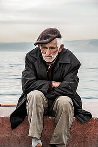 man wearing black leather duffel coat with gray denim jeans and flat cap near bay thumbnail