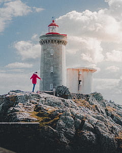 person in red jacket standing on rock formation near lighthouse thumbnail
