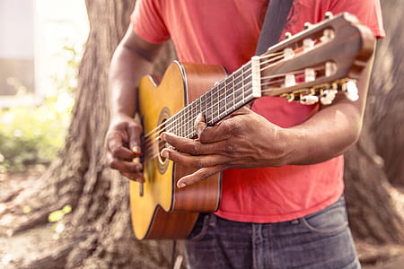 man wearing red t-shirt playing brown classical acoustic guitar near tree thumbnail