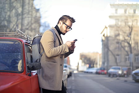 Young Adult bearded male backpacker with backpack on his shoulders, holding phone while leaning on red car thumbnail