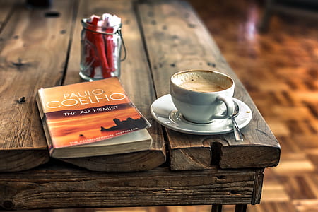 closeup photography of Paulo Coelho The Alchemist book near white ceramic teacup on saucer plate with stainless steel teaspoon on wooden table thumbnail
