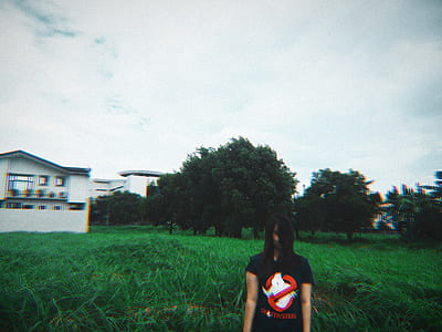 woman wearing black Ghostbuster print tee shirt standing on green field near white building thumbnail