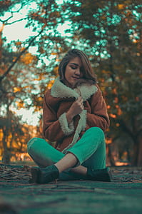 Woman Wearing Brown-and-white Coat and Teal Denim Jeans Sitting on Road Surface thumbnail