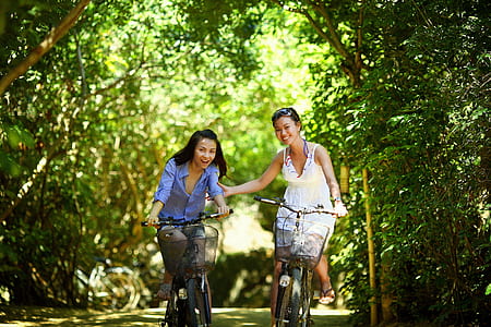 two girl's riding on bicycle passing through green tunnel of trees thumbnail