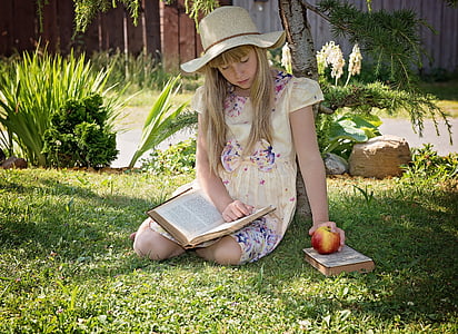 girl wearing dress sitting on grass holding apple and reading book thumbnail