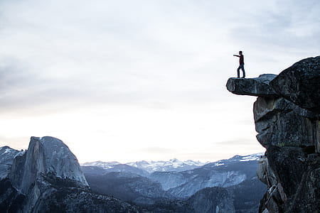 silhouette of person standing on edge of rocky mountain cliff under white cloudy sky thumbnail