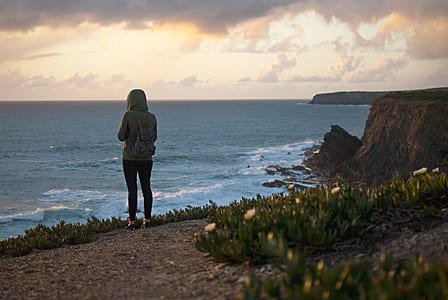 woman standing on cliff facing towards body of water thumbnail