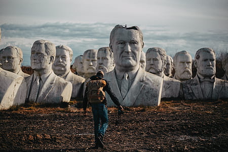 man wearing brown backpack walking in front of concrete human head statues during daytime thumbnail