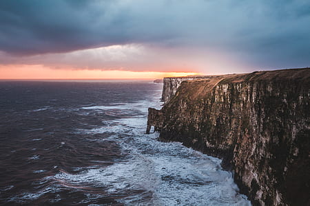 brown cliff near body of water under blue clouds with orange golden time horizon thumbnail