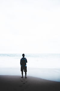 man standing on brown shore near body of water under white sky thumbnail