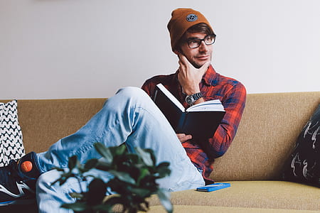 man sitting on sofa holding book and his chin beside wall thumbnail