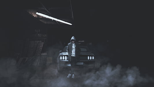 grayscale photography of man wearing hoodie leaning on vehicle bumper thumbnail