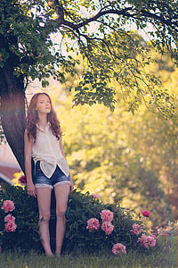 photo of woman in white button-up top and blue denim short shorts standing in front of brown tree and pink flowers thumbnail