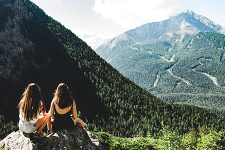 Two Women Sitting on Rock Facing Mountains Under White Cloudy Skies thumbnail