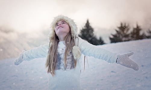 girl wearing fur cap and white crew-neck long-sleeved shirt opening his arms to catch snow during daytime thumbnail