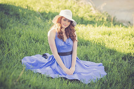 woman wearing blue and white sleeveless polka-dot circle dress and white sun hat sitting on grass field thumbnail