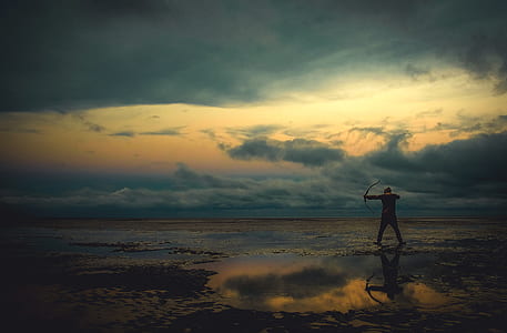 silhouette photo of person holding bow while standing on body of water thumbnail