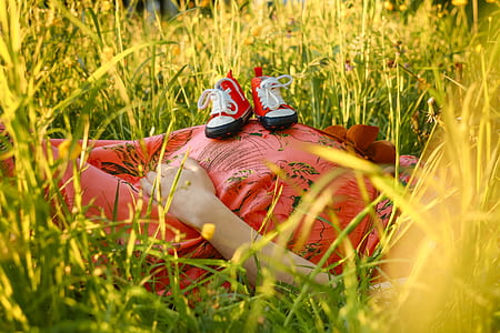 woman wearing red dress lying on grass with toddlers sneakers on her belly thumbnail