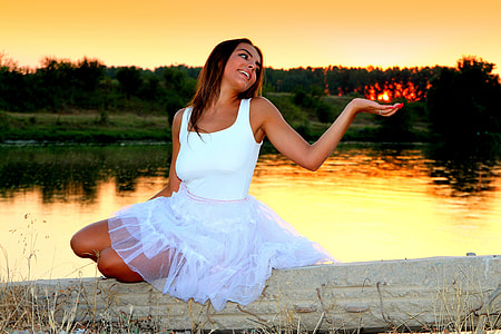 woman in white mini dress sitting on concrete base near body of water during golden hour thumbnail