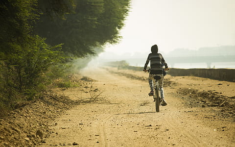 boy riding bicycle on dirt pathway beside body of water during daytime thumbnail