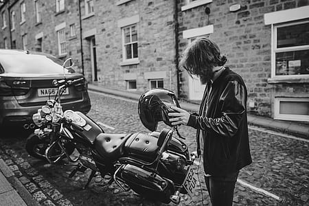 grayscale photo of man standing near motorcycle holding helmet thumbnail