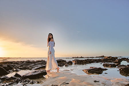 woman in white off-shoulder dress walks on rock over body of water thumbnail